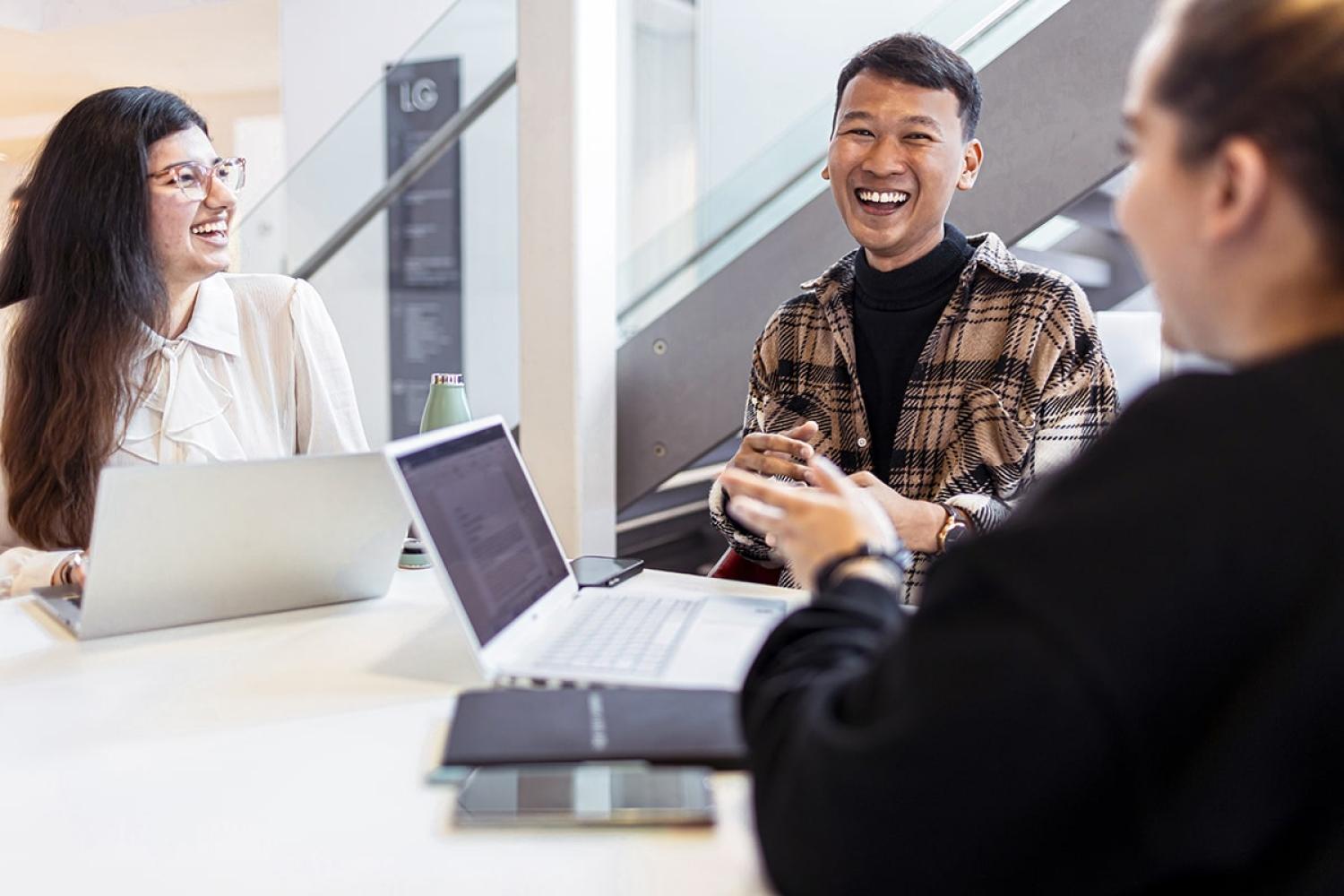 Three smiling students sit around a desk with laptops