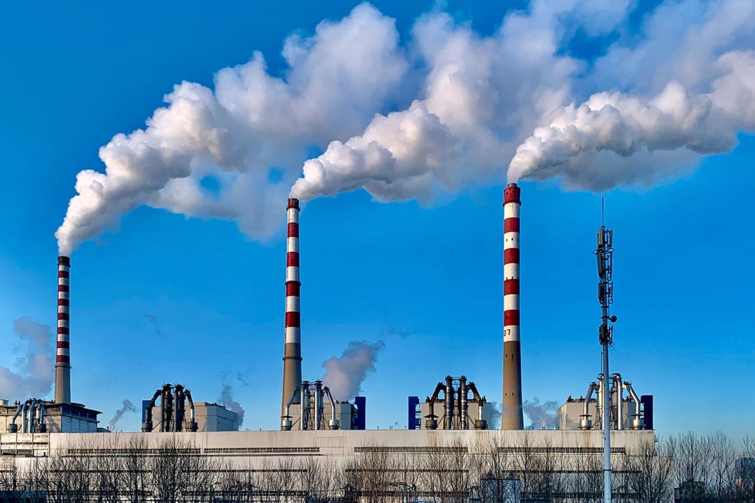 an industrial site with three tall chimneys billowing smoke into a clear blue sky
